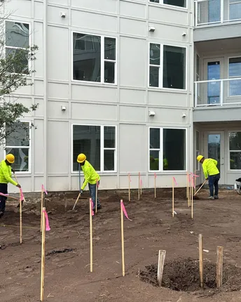 A crew setting grade stakes and moving earth in front of a multi-story residential building.